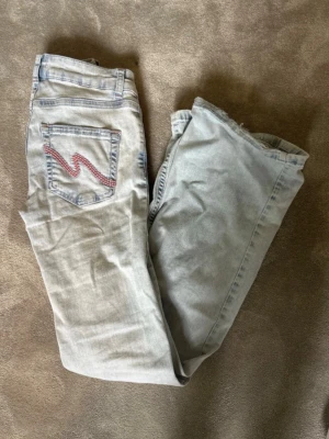 Ljusblå bootcut jeans med slitningar - Säljer ett par ljusblå bootcut jeans med slitningar på båda knäna och en cool broderad detalj i rosa på bakfickan. Jeansen är low waist. Skriv för fler bilder!!!