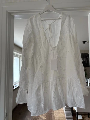 Vit broderad kortklänning med volang - Superfin vit kortklänning med broderade blommor och volangdetaljer. Klänningen har lång ärm, v-ringning med knyt i nacken och en luftig passform. Perfekt för sommarens alla tillfällen.