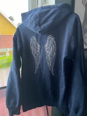 Mörkblå hoodie med strasstenar - Snygg mörkblå hoodie med huva och långa ärmar. På ryggen finns stora vingar i strasstenar och framtill står det 'Ma Chérie' i samma stil. Mjuk och bekväm, perfekt för dig som gillar detaljer som sticker ut.
