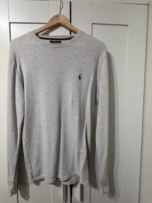 Ljusgrå långärmad tröja Polo Ralph Lauren - Ljusgrå långärmad tröja från Polo Ralph Lauren med diskret svart logga broderad på bröstet. Tröjan har rund halsringning och är tillverkad i ett mjukt, ribbat material som ger en avslappnad och stilren look.