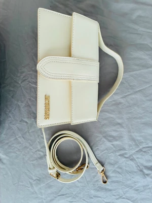 Vit kuvertväska från Jacquemus - Stilren vit kuvertväska från Jacquemus i slätt läder med guldfärgade detaljer och avtagbar axelrem. Väskan har en minimalistisk design med logotypen i guld på framsidan och praktiskt handtag upptill.