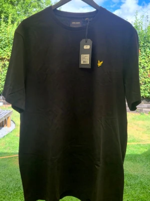 Svart t-shirt från Lyle & Scott - Snygg svart t-shirt från Lyle & Scott med klassisk rund hals och korta ärmar. T-shirten har en gul broderad logga på bröstet och är tillverkad i mjuk bomull. Perfekt för en stilren och enkel look.