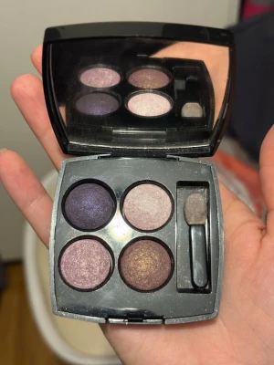 Chanel ögonskuggspalett 4 nyanser - Snygg ögonskuggspalett från Chanel med fyra skimrande nyanser i lila, rosa, beige och brons. Kommer i en elegant svart dosa med spegel och applikator. Perfekt för att skapa olika ögonlooks med både ljusa och mörka toner.