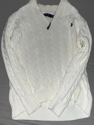 Polo Ralph Lauren Quarter Zip - Kabelstickad vit tröja från Polo Ralph Lauren med halv dragkedja och broderad logga på bröstet. Tröjan har ribbade muddar och hög krage, perfekt för kyliga dagar. Klassisk och tidlös design som passar till det mesta.