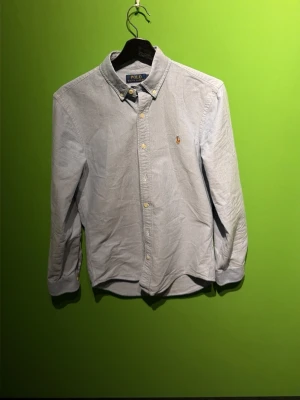Ljusblå skjorta från Polo Ralph Lauren - Klassisk ljusblå skjorta från Polo Ralph Lauren med broderad logga på bröstet. Skjortan har knappar framtill, button-down krage och långa ärmar. Perfekt till jeans eller chinos för en clean och stilren look.