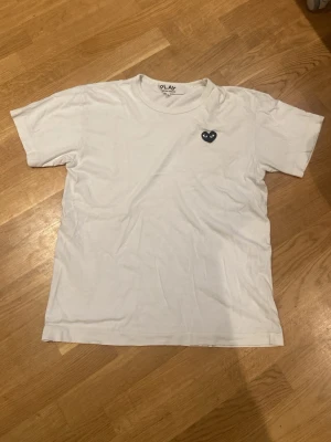 Vit Comme des Garçons PLAY t-shirt - Vit t-shirt från Comme des Garçons PLAY med det ikoniska svarta hjärta-trycket på bröstet. Klassisk rund halsringning och korta ärmar. Tillverkad i mjuk bomull för en skön och avslappnad look. Är medium men sitter som en small.