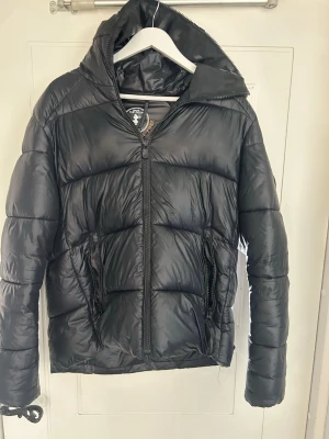 Svart Save The Duck jacka - Säljer en svart pufferjacka med märket Save The Duck med huva och dragkedja framtill. Materialet är glansigt och jackan är perfekt för kalla vinterdagar. Men som ni ser på bilderna finns några skavanker som är ihopsydda, kostar ca 3000 ny.