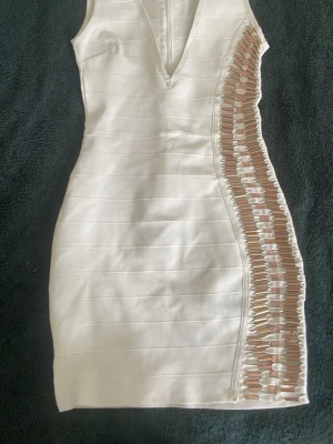 Vit kort klänning med metall detaljer - Snygg vit kort klänning med v-ringning och unik metalldetaljer längs sidan som ger en cool look. Klänningen är ärmlös och har en figurnära passform. Perfekt för dig som vill sticka ut med en trendig och modern stil. Den är i 90% polyester och 10% elastisk material.