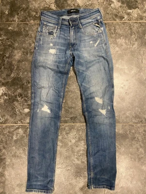 Blå slitna jeans från Replay - Säljer ett par ljusblå jeans från Replay med fit och slitna detaljer på både fram- och baksida. Jeansen har klassisk femficksmodell, dragkedja och knapp i midjan samt Replay-lapp bak i midjan. Perfekta för en avslappnad och trendig look.