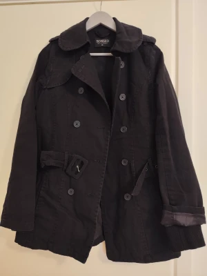 Svart trenchcoat från Soaked in Luxury - Stilren svart trenchcoat med dubbelknäppning, bred krage och bälte i midjan. Jackan har ribbade muddar vid ärmsluten och detaljer i satin på ärmarna. Tillverkad i ett kraftigare tyg som ger en snygg siluett. Perfekt för dig som gillar klassisk men modern stil.
