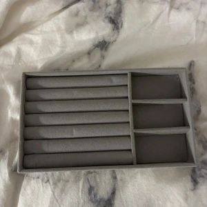 Grå smyckesförvaring i sammet - Snygg och praktisk smyckesförvaring i grå sammet med flera fack och rader för ringar, örhängen eller andra småsmycken. Perfekt för att organisera dina accessoarer på ett stilrent sätt.