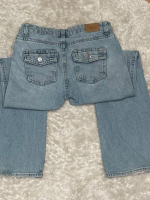 Ljusblå bootcut jeans med fickor - Säljer ett par ljusblå jeans med bootcut passform och två stora fickor med lock och knapp bak. Jeansen har klassisk femficksdesign fram och är tillverkade i denim. Perfekta för en avslappnad och trendig look.