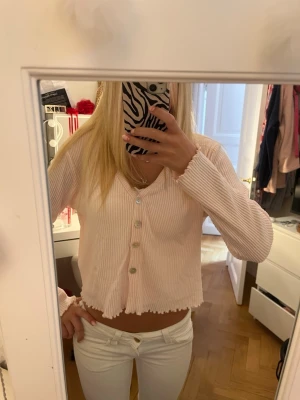 Ljusrosa ribbad kofta med knappar - Supersöt ljusrosa ribbad kofta med v-ringning och fyra knappar framtill. Modellen är croppad med vågig kant nertill och långa ärmar. Perfekt att slänga över ett linne eller topp för en chill look. Jag har köpt den på Newyorker men har inte andvänt den. Storleken på koftan är L men den passar som S💗