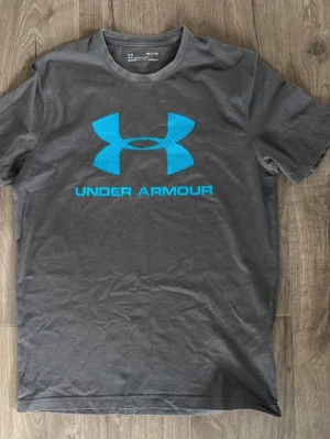 Grå t-shirt från Under Armour - Snygg grå t-shirt från Under Armour med stort blått logotryck på bröstet. Klassisk passform med rund halsringning och korta ärmar. Perfekt för dig som gillar sportig stil och vill ha något bekvämt till vardags.