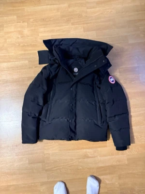 Svart pufferjacka från Canada Goose - Säljer en svart pufferjacka från Canada Goose med klassisk patch på ärmen. Jackan har stor huva, dragkedja och knappar framtill samt två sidofickor. Perfekt för kalla vinterdagar och riktigt varm tack vare dunfyllning.