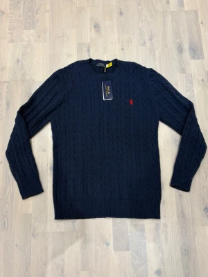 Mörkblå kabelstickad tröja Polo Ralph Lauren - Snygg mörkblå kabelstickad tröja från Polo Ralph Lauren med rund halsringning och den klassiska röda logotypen broderad på bröstet. Tröjan har ribbade muddar vid ärmslut och nederkant. Perfekt för dig som gillar stilrena och tidlösa plagg.
