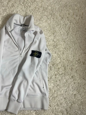 Vit zip hoodie från Stone Island - Säljer en clean vit hoodie från Stone Island med dragkedja framtill och två stora fickor. Klassisk logopatch på ärmen och ribbade muddar. Tillverkad i mjukt bomullsmaterial för skön känsla. Perfekt för dig som  vill ha något stilrent.