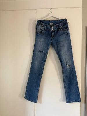 Blå slitna jeans från LTB - Säljer ett par blå jeans från LTB med slitningar och hål på båda benen. Jeansen har raka ben, klassisk femficksmodell och knappar i gylfen. Perfekta för en avslappnad och trendig look.