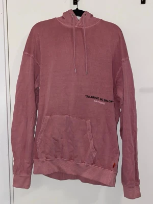 Rosa hoodie med tryck och huva - Säljer en oversized rosa hoodie med huva och dragsnören. Framtill finns en stor magficka och ett svart tryck med texten 'AS ABOVE, SO BELOW' samt 'VLNT©2002'. Tillverkad i mjukt bomullstyg och har ribbade muddar vid ärmar och nederkant.