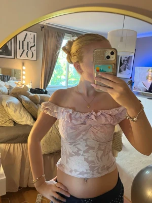 Ljusrosa offshoulder blus med volang - Supersöt ljusrosa offshoulder blus med volangkant och subtilt blommönster. Toppen är croppad och har en romantisk känsla med rynkdetalj framtill. Perfekt för dig som vill ha en feminin och trendig look.