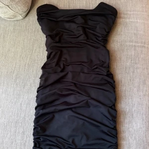 Svart axelbandslös tight klänning - Säljer en svart axelbandslös klänning med tight passform och rynkade detaljer längs hela klänningen. Klänningen är kort och har en elegant, figurnära siluett som framhäver formerna. Perfekt för dig som vill ha en stilren och trendig look.