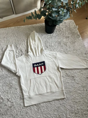Gant Vit hoodie - Snygg vit hoodie från GANT med stor broderad logga och sköld på bröstet i rött, vitt och blått. Klassisk känguruficka framtill och ribbade muddar vid ärmar och nederkant. Tillverkad i mjukt bomullsmaterial och har en stor huva. Skriv till mig för fler frågor.🙌 11-12 år