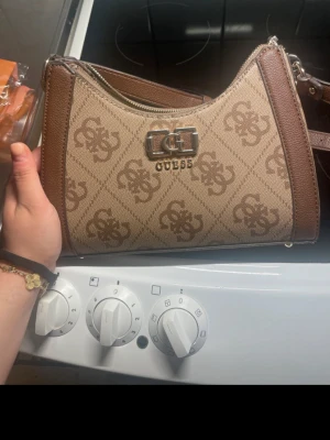Beige och brun axelväska från Guess - Snygg axelväska från Guess i beige och brunt med logomönster över hela väskan. Väskan har silverfärgad metallogo framtill, dragkedja upptill och justerbar axelrem. Insidan är fodrad med Guess-tryck och har en innerficka med dragkedja. Perfekt för att lyfta din outfit.