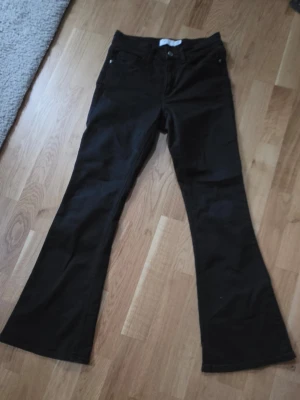 Svarta bootcut jeans - Säljer ett par svarta bootcut jeans med klassisk femficksdesign och utsvängda ben. Jeansen har knapp- och dragkedjestängning framtill och är sydda i ett mjukt denimtyg. Perfekta för dig som gillar retrovibbar och vill ha en snygg siluett.
