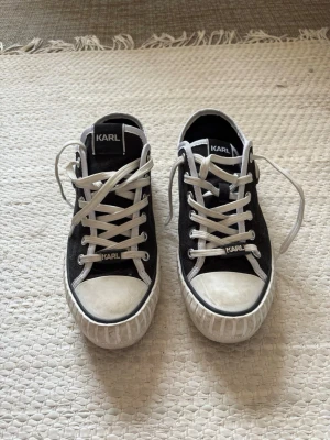 Svarta Karl Lagerfeld sneakers - Snygga svarta sneakers från Karl Lagerfeld med vita detaljer och vita snören. Skorna har en ikonisk Karl Lagerfeld-logga på sidan och en robust vit sula med mönster. Klassisk låg modell som passar till många olika stilar.