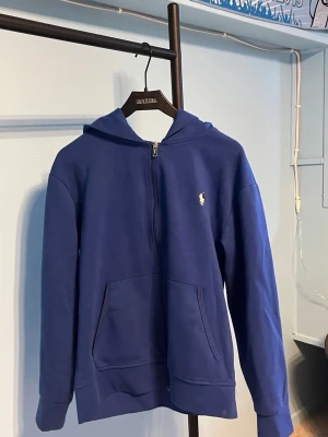 Blå hoodie med dragkedja och huva - Snygg blå hoodie med dragkedja framtill och klassisk huva. Har en liten vit logga broderad på bröstet och stora fickor framtill. Tillverkad i mjukt bomullsmaterial som känns skönt mot huden. Perfekt för en avslappnad och stilren look.