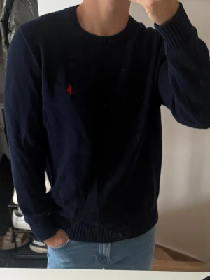 Mörkblå stickad tröja från Polo Ralph Lauren - Mörkblå stickad tröja från Polo Ralph Lauren med klassisk röd logga broderad på bröstet. Tröjan har rund halsringning, långa ärmar och ribbstickade muddar vid ärmslut och nederkant. Perfekt till jeans för en clean look.