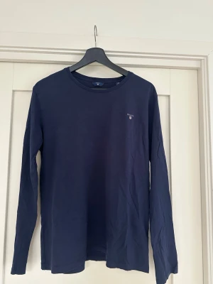 Mörkblå långärmad T-shirt  från Gant - En stilren mörkblå långärmad topp från Gant med rund halsringning och diskret logga på bröstet. Tillverkad i mjuk bomull som känns skön mot huden. Perfekt att matcha med jeans eller kjol för en clean look.