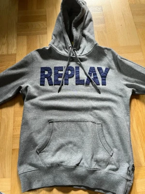 Grå hoodie från Replay med tryck - Snygg grå hoodie från Replay med stort blått REPLAY-tryck på bröstet. Klassisk känguruficka framtill och justerbar huva med dragsko. Mjuk bomullsblandning som är skön att ha på sig. Perfekt för en avslappnad look. Lite sliten på fikan och öglan man hänger upp den med har lossnat 