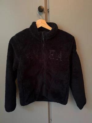 Svart teddyjacka från SVEA - Mysig svart teddyjacka från SVEA med hög krage och dragkedja framtill. Jackan har broderad logga på bröstet och är gjord i fluffigt, mjukt material. Perfekt för kyliga dagar när du vill vara både varm och stilren.