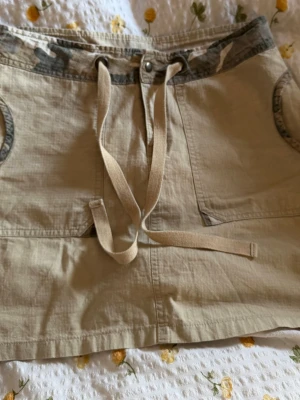 Beige kortkjol med camo-detaljer - Snygg beige kortkjol med camouflagemönstrade detaljer i midjan och på fickorna. Kjolen har stora framfickor, bakfickor och snörning framtill. Perfekt för en avslappnad och cool stil.