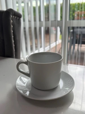Vit kaffekopp med fat 9X  - En stilren vit kaffekopp i porslin med matchande fat. Koppen har ett klassiskt handtag och rund form, perfekt för kaffe eller te. Enkel och tidlös design som passar alla tillfällen.