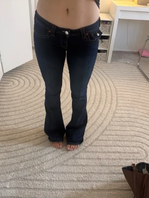 Mörkblå bootcut jeans True Religion - Säljer ett par mörkblå bootcut jeans från True Religion med låg midja och kontrastsömmar. Jeansen har klassiska fickor fram och bak, samt den ikoniska loggan och detaljerade sömmar på bakfickorna. Perfekta för en avslappnad men snygg look. Använda 2 gånger, jag bär storlek 36/38 och dom passar bra, stretchiga 