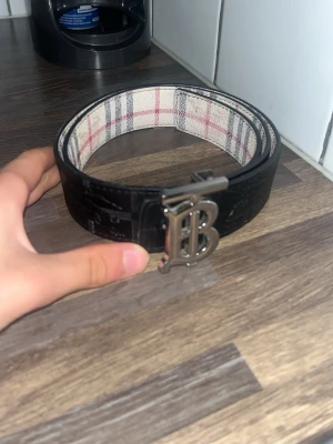 Burrberry bälte  - Snyggt svart bälte med ett stort B-format metallspänne. Insidan har ett rutigt mönster i vitt, rött och grått. Perfekt accessoar för att lyfta din outfit.