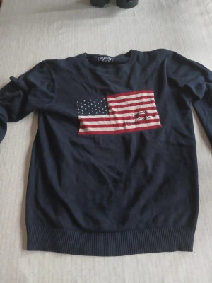 Mörkblå tröja med USA-flagga Vinson - Mörkblå långärmad tröja från Vinson Polo Club med stor amerikansk flagga broderad på bröstet. Tillverkad i 100% bomull, rund hals och ribbade muddar vid ärmslut och nederkant. Perfekt för dig som gillar klassisk och sportig stil.