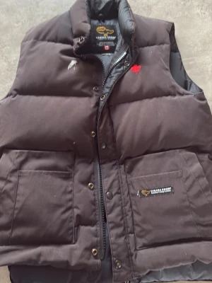 Svart dunväst från Canada Goose - Snygg svart dunväst från Canada Goose med hög krage och två stora fickor framtill. Västen har en röd lönnlogga på bröstet och klassisk Canada Goose-märkning. Perfekt för lager-på-lager och håller dig varm under kyliga dagar.