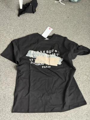 Maison Margiela svart t-shirt - Cool svart t-shirt från Maison Margiela med tryck framtill i vitt och beige samt logga. Klassisk passform med korta ärmar och rund hals. Perfekt för dig som gillar streetwear och vill ha något unikt i garderoben.