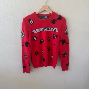 Röd stickad tröja Love Moschino - Röd stickad tröja från Love Moschino med svarta och vita grafiska tryck som hjärtan, stjärnor och läppstift. Rund halsringning och långa ärmar. Perfekt för dig som vill sticka ut med en färgstark och lekfull stil.0233