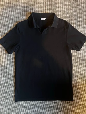 Svart pikétröja från Filippa K - Klassisk svart pikétröja från Filippa K i storlek M. Tröjan har korta ärmar, krage och knapplös öppning framtill. Perfekt till både jeans och chinos för en clean och stilren look. Nypris 900kr
