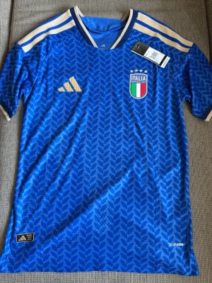 Blå Italien fotbollströja Adidas - Snygg blå fotbollströja från Adidas med Italiens landslagsmärke på bröstet och guldiga detaljer på axlar och logga. Tröjan har ett mönster över hela tyget och korta ärmar. Perfekt för dig som älskar fotboll och vill stötta Italien.