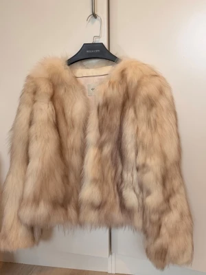 Beige pälsjacka från Coco Limited - Säljer en fluffig och trendig beige pälsjacka från Coco Limited. Jackan har en mjuk och lyxig känsla med långa ärmar och utan krage. Perfekt för dig som vill sticka ut med en statement piece i garderoben.