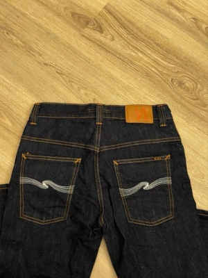 Mörkblå nudie jeabs  - Säljer ett par mörkblå nudie jeans med raka ben och snygga kontrastsömmar. Bakfickorna har en vit vågig brodyr och läderpatch i midjan. Klassisk femficksmodell i kraftig denim, perfekt till vardagslooken. Pris går att diskutera 