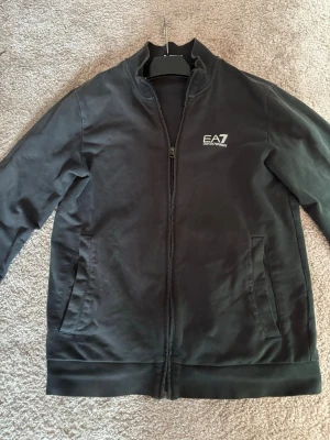 Svart zip-hoodie från EA7 Emporio Armani - Snygg svart hoodie med dragkedja från EA7 Emporio Armani. Hoodien har två fickor framtill och en diskret logga på bröstet. Tillverkad i mjukt material som känns skönt mot huden. Perfekt för en avslappnad och stilren look.