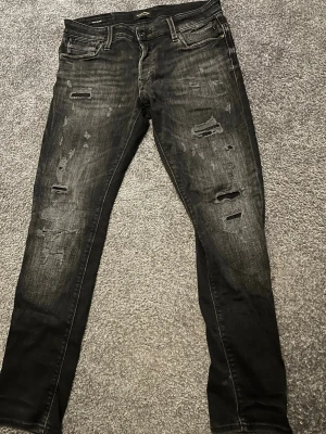 Svarta slitna jeans från Jack & Jones - Säljer ett par svarta jeans från Jack & Jones med slitna detaljer och slim fit. Jeansen har klassisk femficksmodell, dragkedjegylf och snygga slitningar framtill för en trendig look.