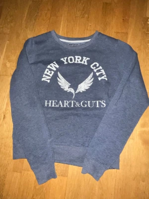 Blå sweatshirt Heart & Guts S - Mysig blå sweatshirt från Heart & Guts med tryck 'New York City' och vingar på bröstet. Tröjan har rund halsringning, långa ärmar och mjukt material på insidan. Perfekt för en avslappnad och cool stil.