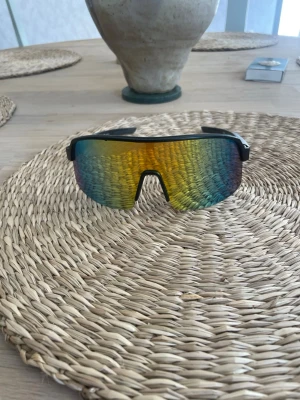 Svarta sportiga solglasögon - Säljer ett par sportiga solglasögon med svart båge och spegelglas i regnbågsfärger. Glasen har en bred, svepande form som ger en futuristisk look och passar perfekt för en aktiv livsstil. Snygga att bära till både streetwear och sportoutfits.